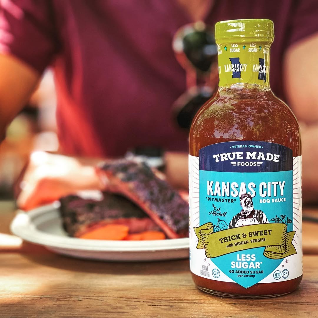 True Made Foods Kansas City BBQ Sauce bottle on a table with blurred food in the background.