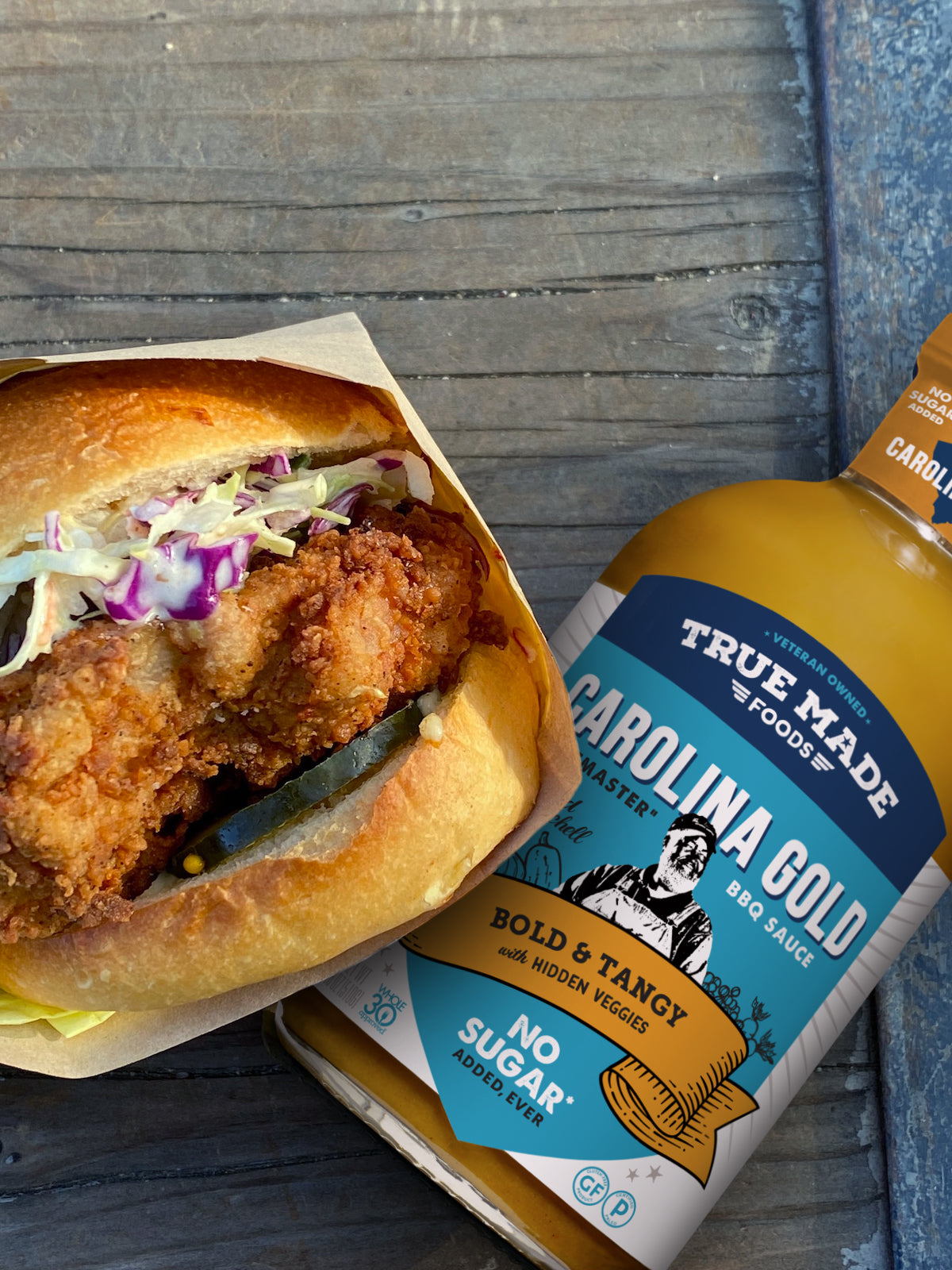A crispy chicken sandwich with coleslaw and pickles, alongside a bottle of “True Made Carolina Gold BBQ Sauce.”.