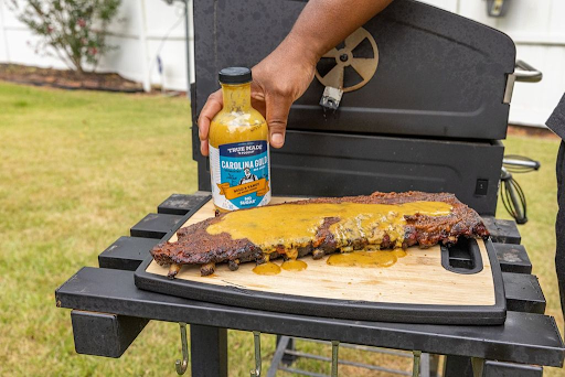 A bottle of “True Made Foods Carolina Gold BBQ Sauce” placed over ribs on a wooden board, next to a grill.