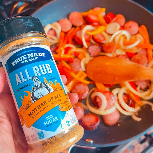 Hand holding True Made Foods All Rub near a pan with cooking sausage, onions, and peppers.