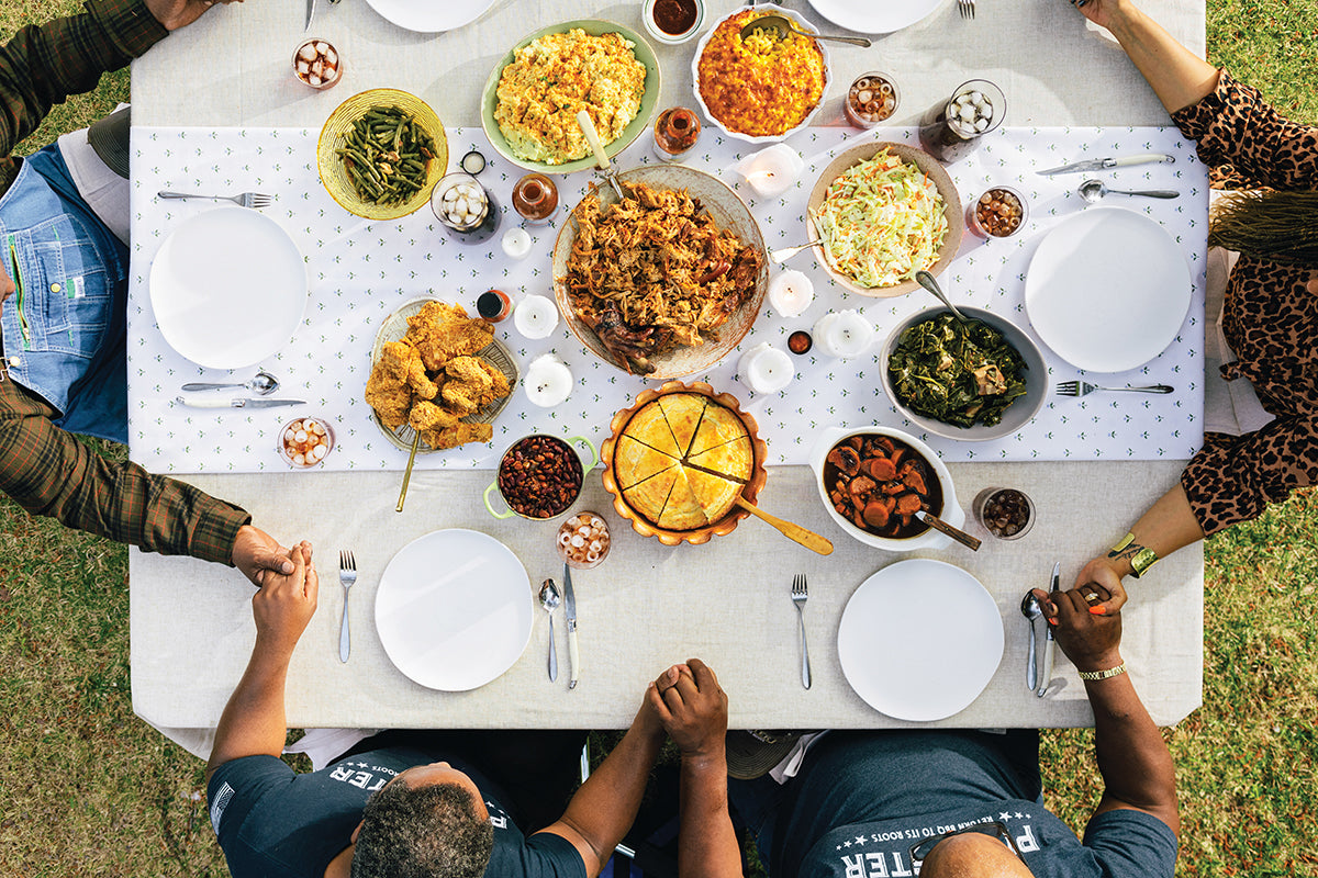 Overhead view of diverse hands joined around a table filled with assorted dishes and drinks, symbolizing shared meals.