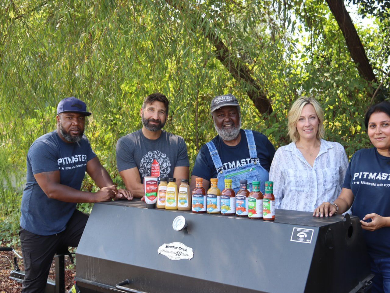 Five people, including three men and two women, pose with True Made Foods BBQ sauce bottles and a grill outdoors.
