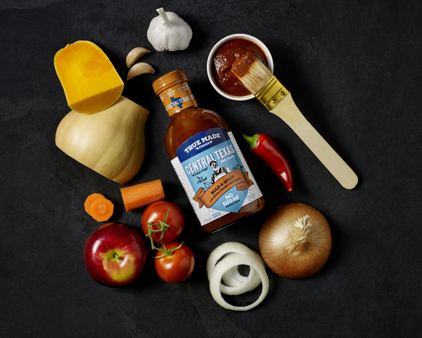  True Made Foods’ Central Texas BBQ sauce bottle surrounded by ingredients like onions, carrots, tomatoes, and red chili.