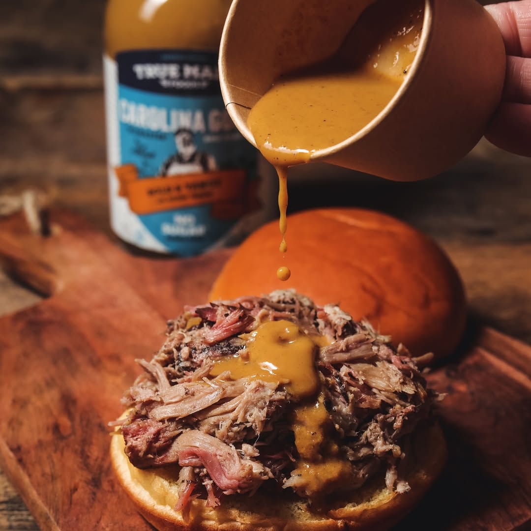 Hand pouring True Made Foods Carolina Gold BBQ sauce onto a pulled-pork sandwich on a wooden board.