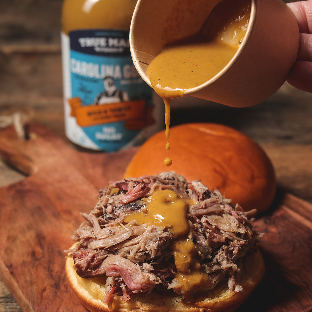 Person pouring True Made Foods sugar-free Carolina mustard BBQ sauce onto a burger.