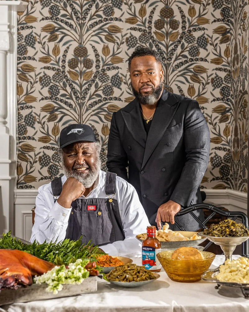Pitmasters Ed and Ryan Mitchell seated at a table with traditional southern barbecue and True Made Foods BBQ sauce.
