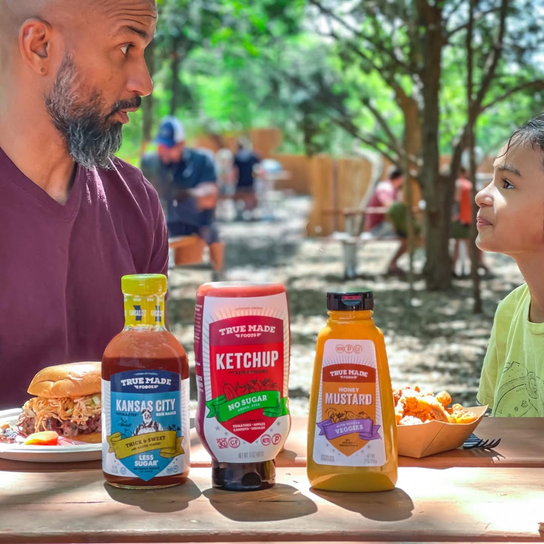 Father and child enjoying burgers outdoors with True Made Foods BBQ sauce, ketchup, and mustard on the table.