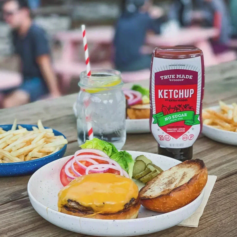 Bottle of True Made Foods No Sugar Ketchup served with a cheeseburger, crispy fries, and a glass of lemonade on a picnic table.