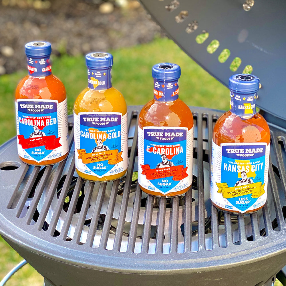 Four bottles of True Made Foods BBQ sauces—Carolina Red, Carolina Gold, E. Carolina, and Kansas City—displayed on a grill.