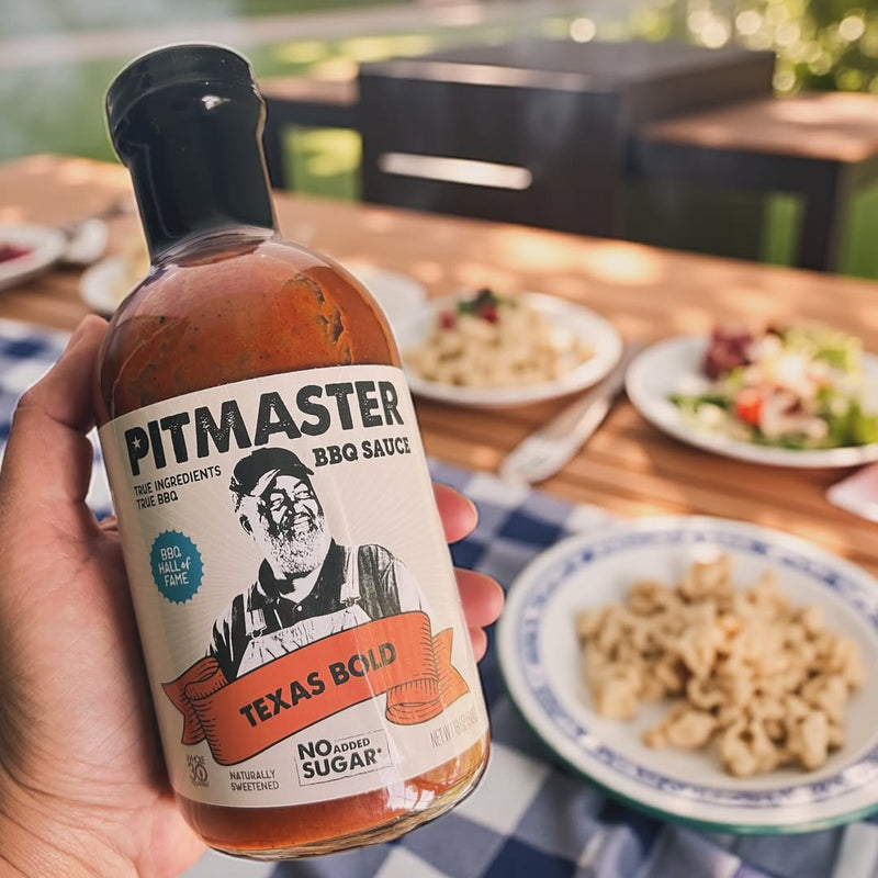 Hand holding True Made Foods Texas Bold BBQ sauce bottle with a meal on the table in the background.