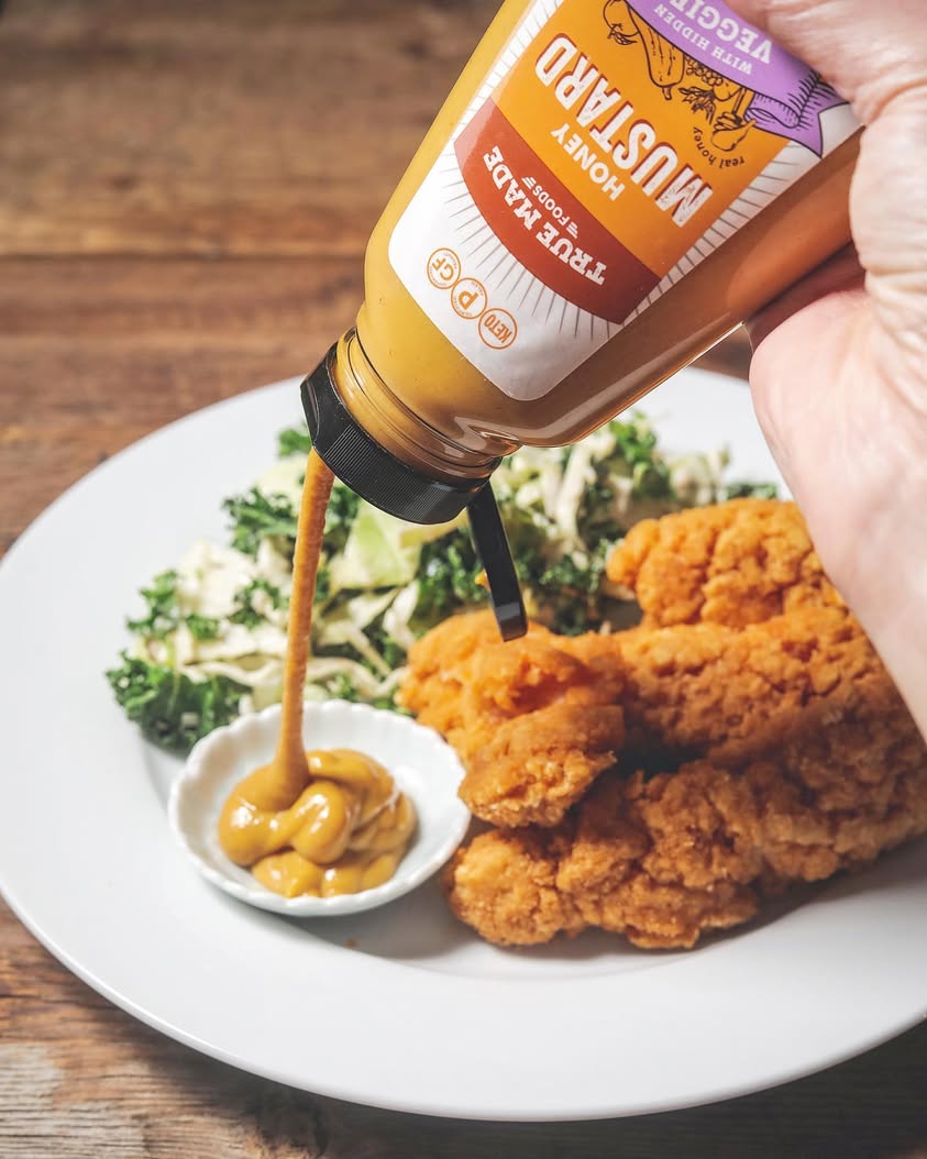 Hand squeezing True Made Foods Honey Mustard into a dipping bowl beside crispy chicken nuggets on a plate.