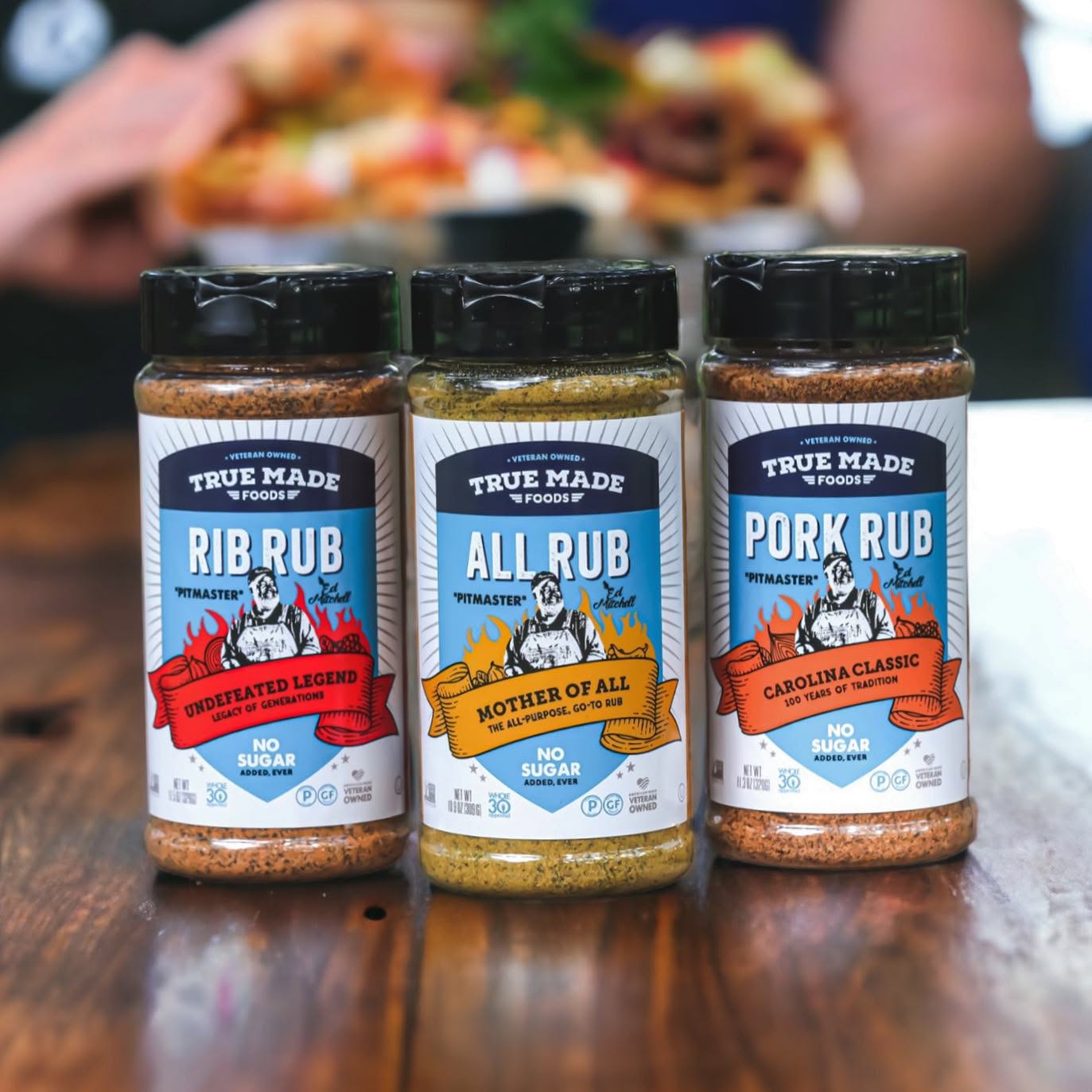 Three jars of True Made Foods BBQ seasoning rubs, including Rib Rub, All Rub, and Pork Rub, displayed on a wooden surface.