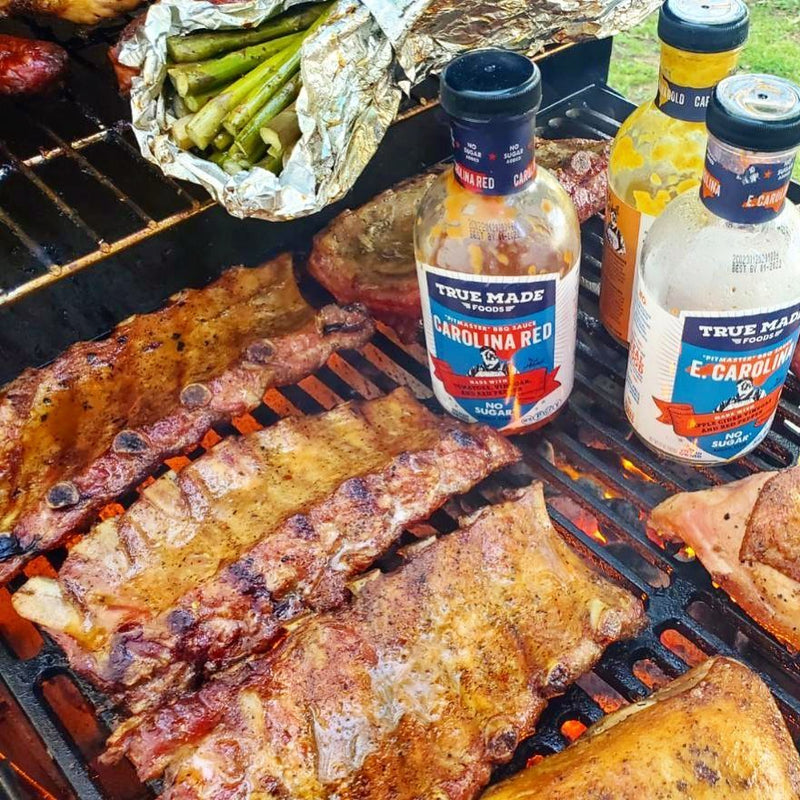 Grilled ribs, chicken, and asparagus cooking over open flames beside bottles of True Made Foods BBQ sauces at a backyard cookout.