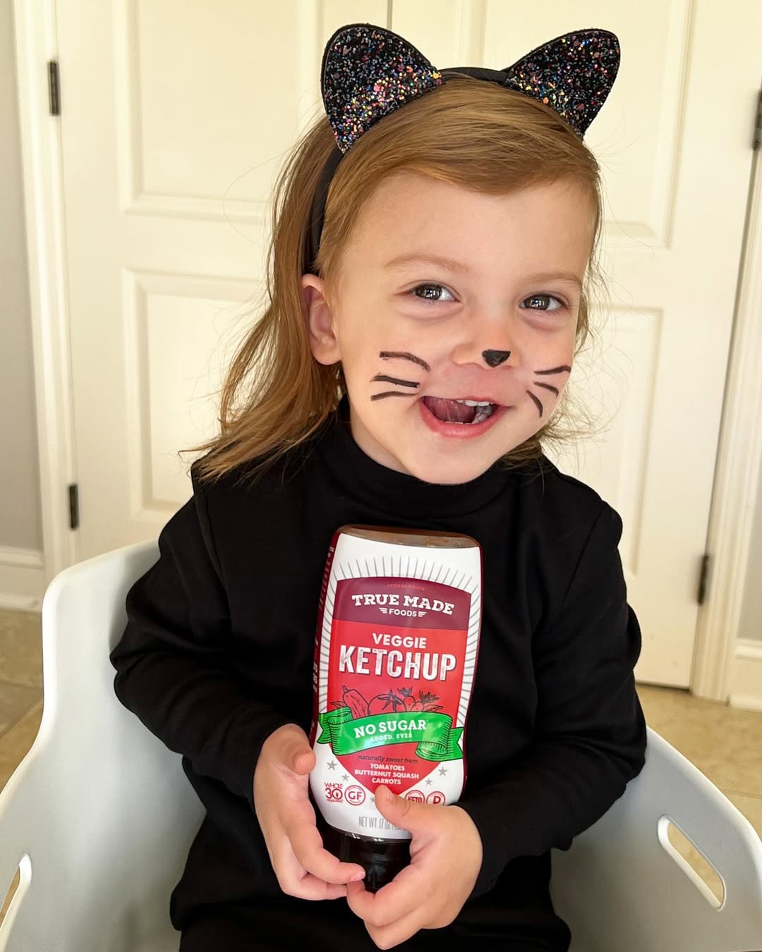 A smiling child in a black cat Halloween costume holding a bottle of True Made Foods veggie ketchup, ready to enjoy healthy Halloween snacks.