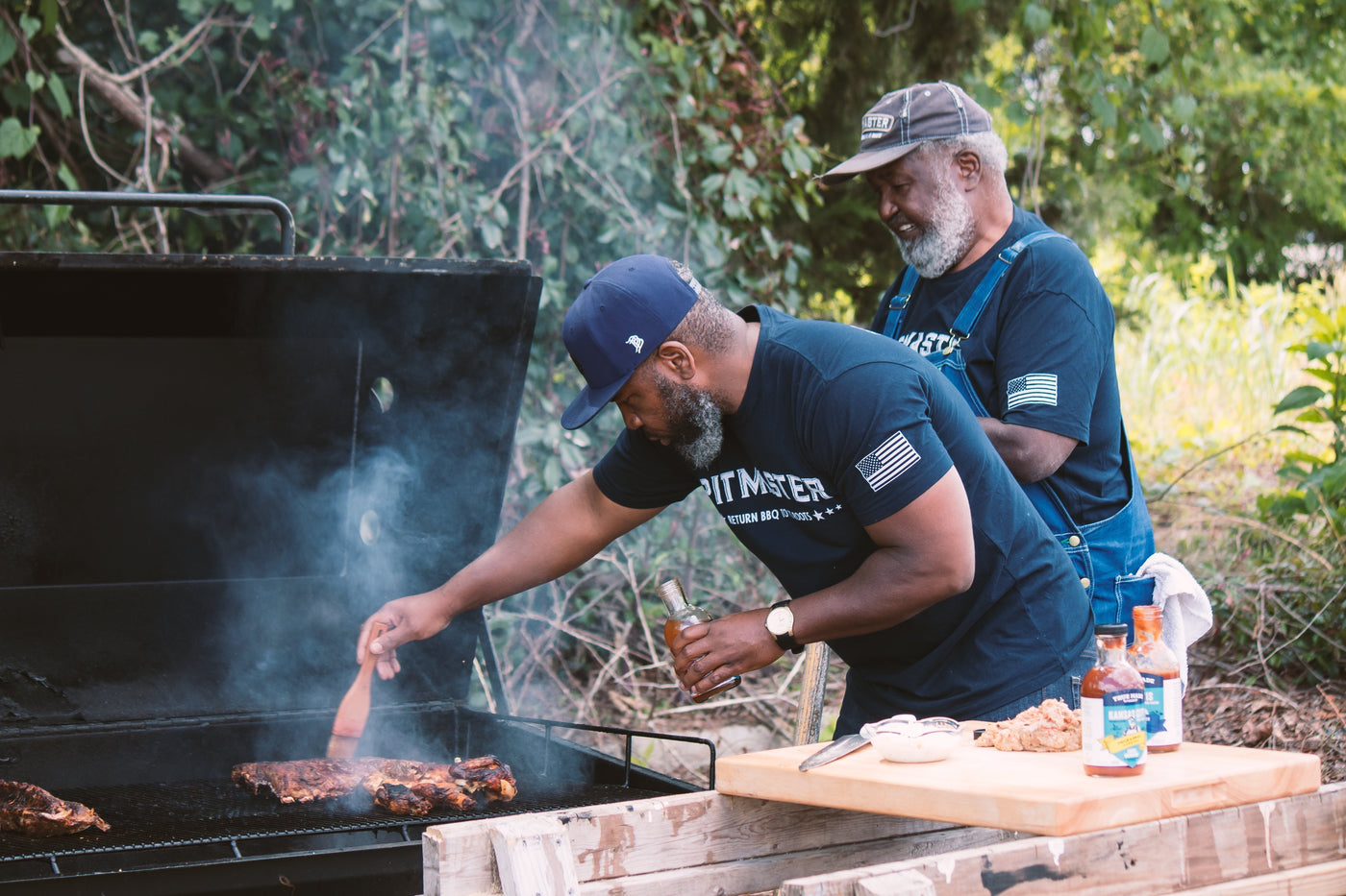 Ed Mitchell and Ryan Mitchell barbecuing in their yard, brushing True Made Foods sugar-free BBQ sauce onto meat.