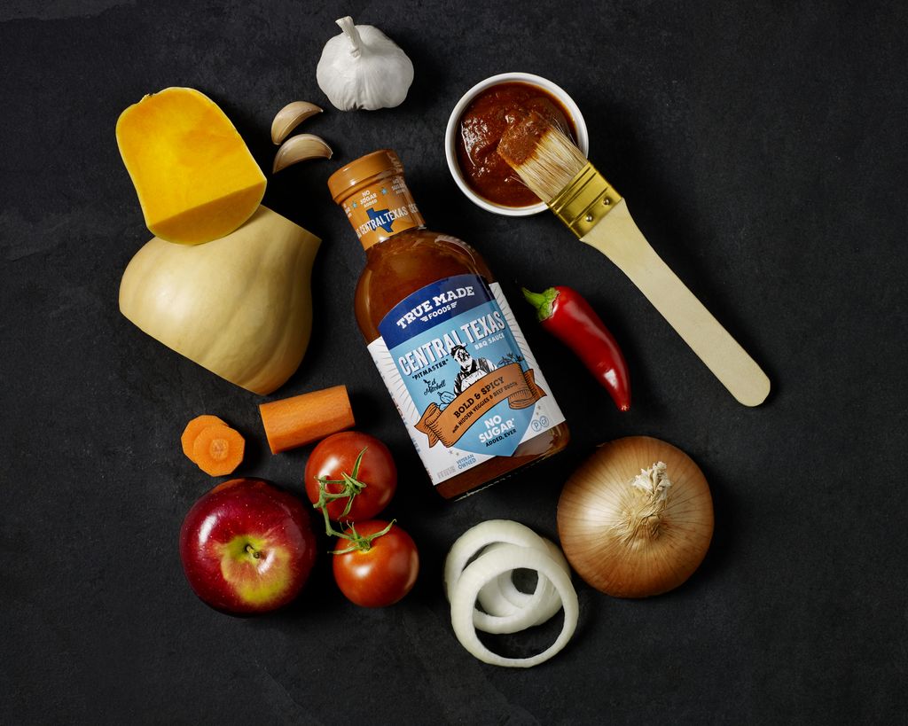 An assortment of fresh vegetables and fruit, a bottle of True Made Foods Central Texas BBQ Sauce, and a basting brush.