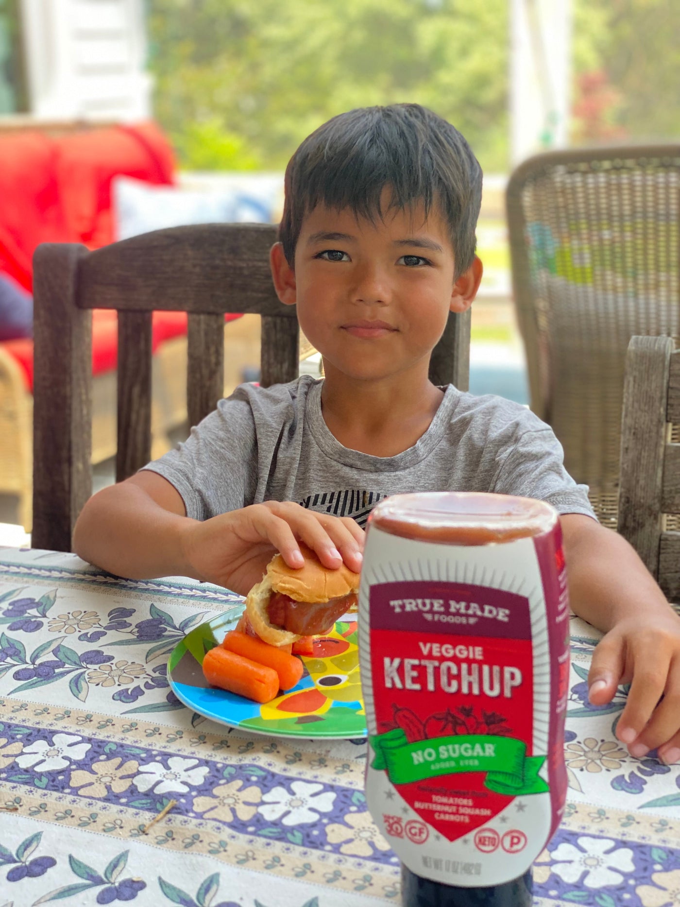 A young boy smiles at a table with a hot dog, carrots, and a ‘True Made Foods Veggie Ketchup’ bottle.