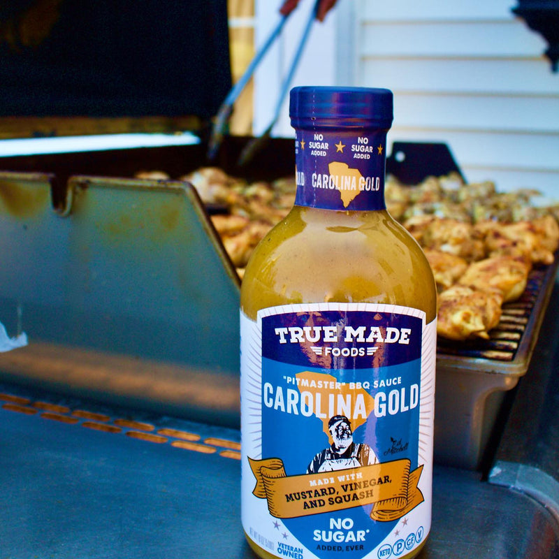 True Made Foods’ Carolina Gold No Sugar BBQ Sauce bottle in front of an outdoor grill with cooking chicken.