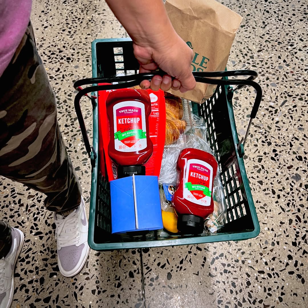 A person holding a grocery basket with a True Made Foods ketchup and bread in it.