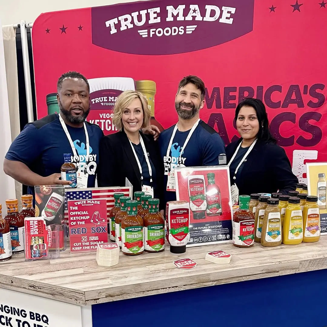 True Made Foods booth at an event with BBQ sauces, hot sauces, ketchup, and mustards displayed on a branded table.