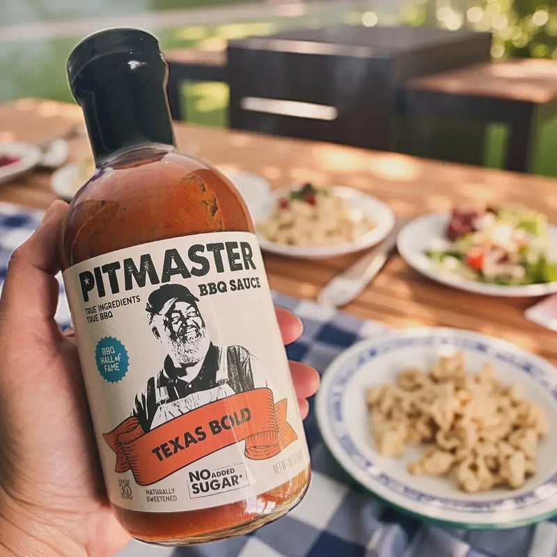 Hand holding a bottle of True Made Foods Texas Bold BBQ Sauce beside grilled food at an outdoor barbecue.