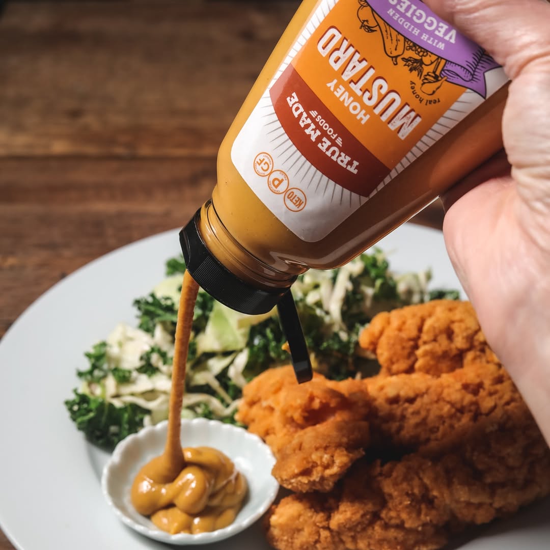 Squeezing True Made Foods Honey Mustard into a dish beside fried chicken and greens on a dinner plate.