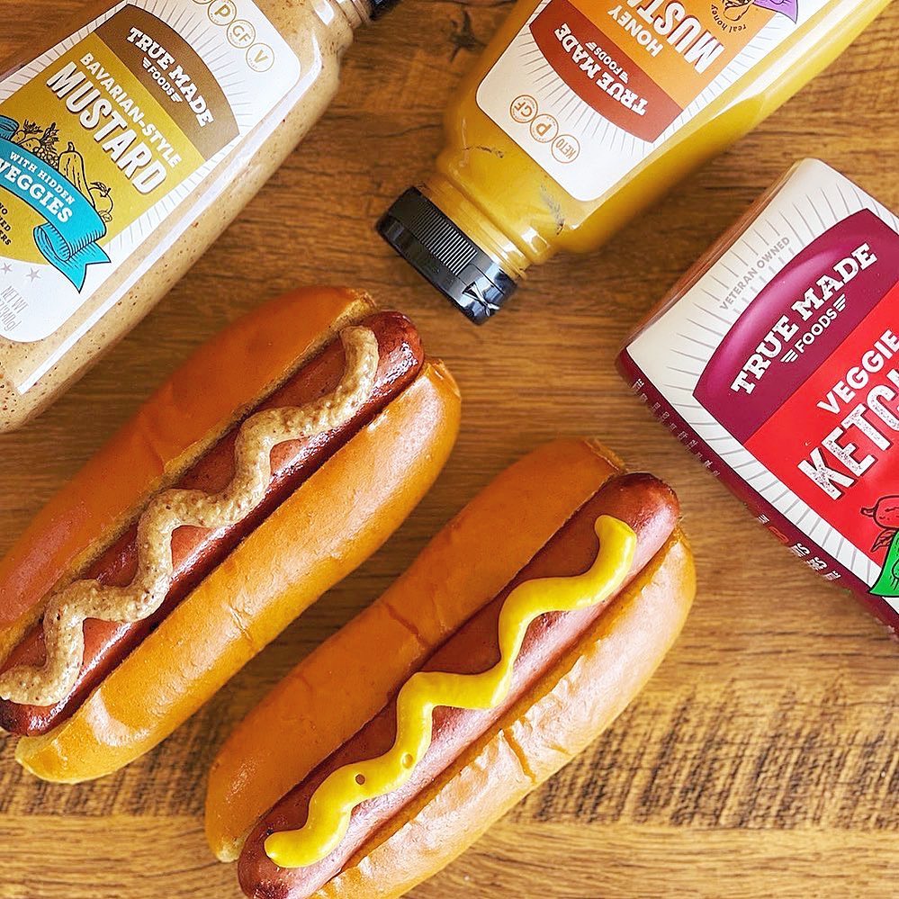 Two hot dogs topped with mustard, with True Made Foods Bavarian Mustard and Ketchup bottles placed beside them.