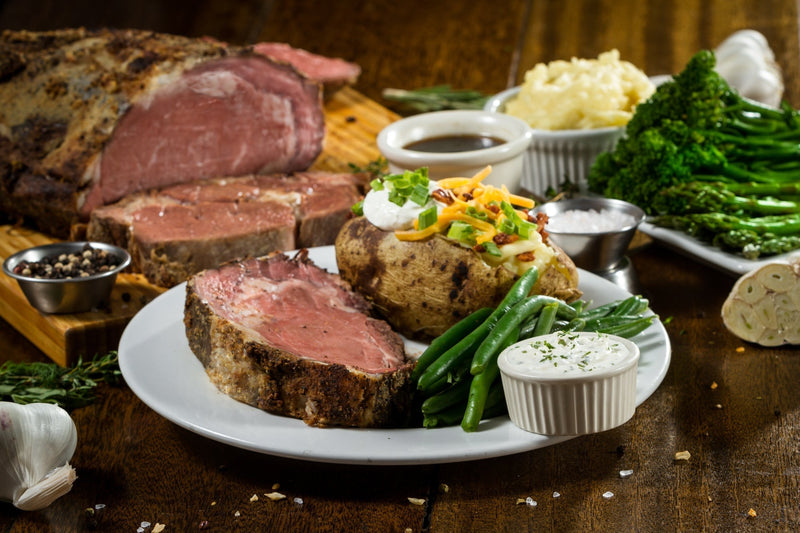 A plate of prime rib roast with a loaded baked potato, green beans, and horseradish sauce on a wooden table.