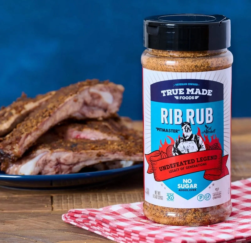 A bottle of True Made Foods Rib Rub sitting next to a plate of seasoned beef ribs.