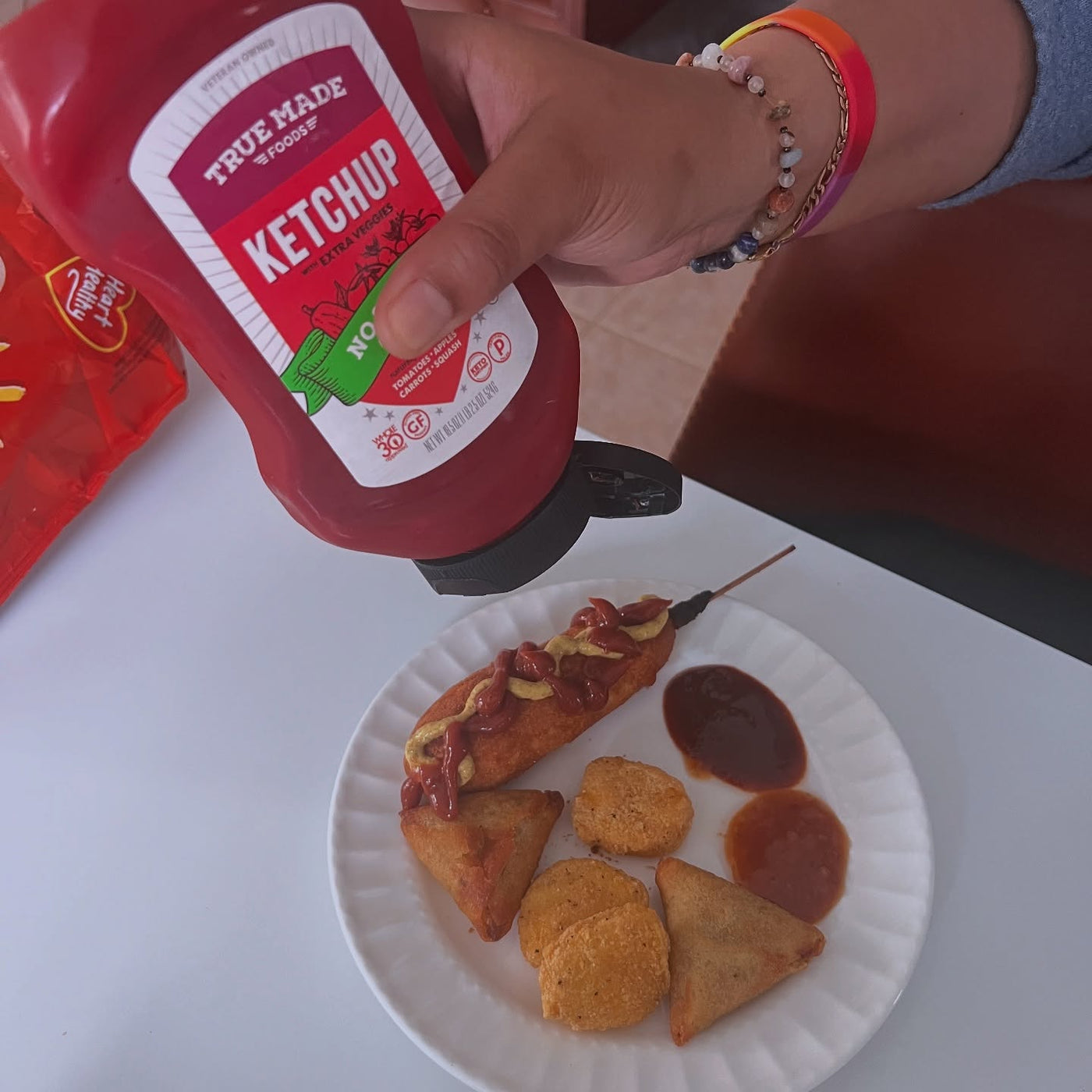 A person squeezing ketchup on a hot dog with assorted fried sauce and dipping sauce from True Made Foods.