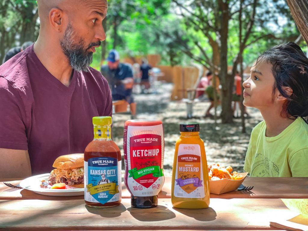 A man sitting with a young girl at a table with food and True Made Foods Kansas City BBQ sauce, ketchup, and mustard.