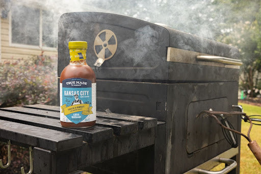 Bottle of True Made Foods BBQ sauce next to a smoking charcoal grill, perfect for a clean-eating BBQ.
