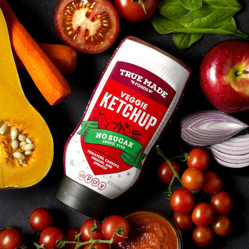 Bottle of True Made Foods Veggie Ketchup surrounded by tomatoes, carrots, squash, and apple, showcasing a no-sugar condiment alternative.