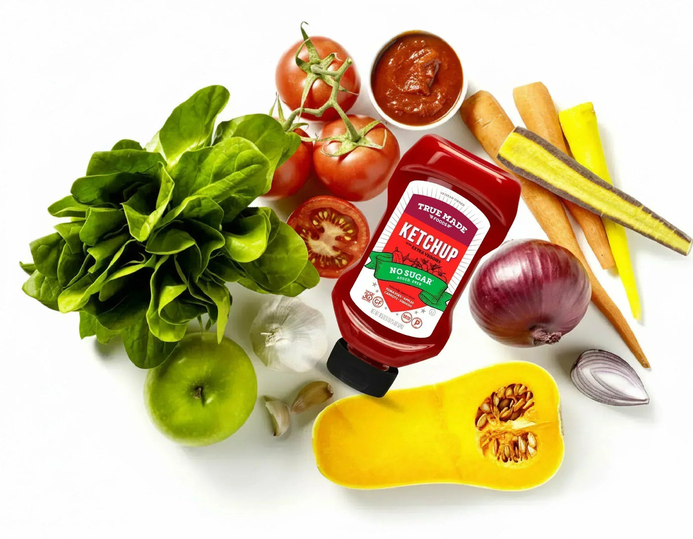 Bottle of True Made Foods No Sugar Veggie Ketchup surrounded by fresh vegetables, including carrots, tomatoes, leafy greens, and a green apple.