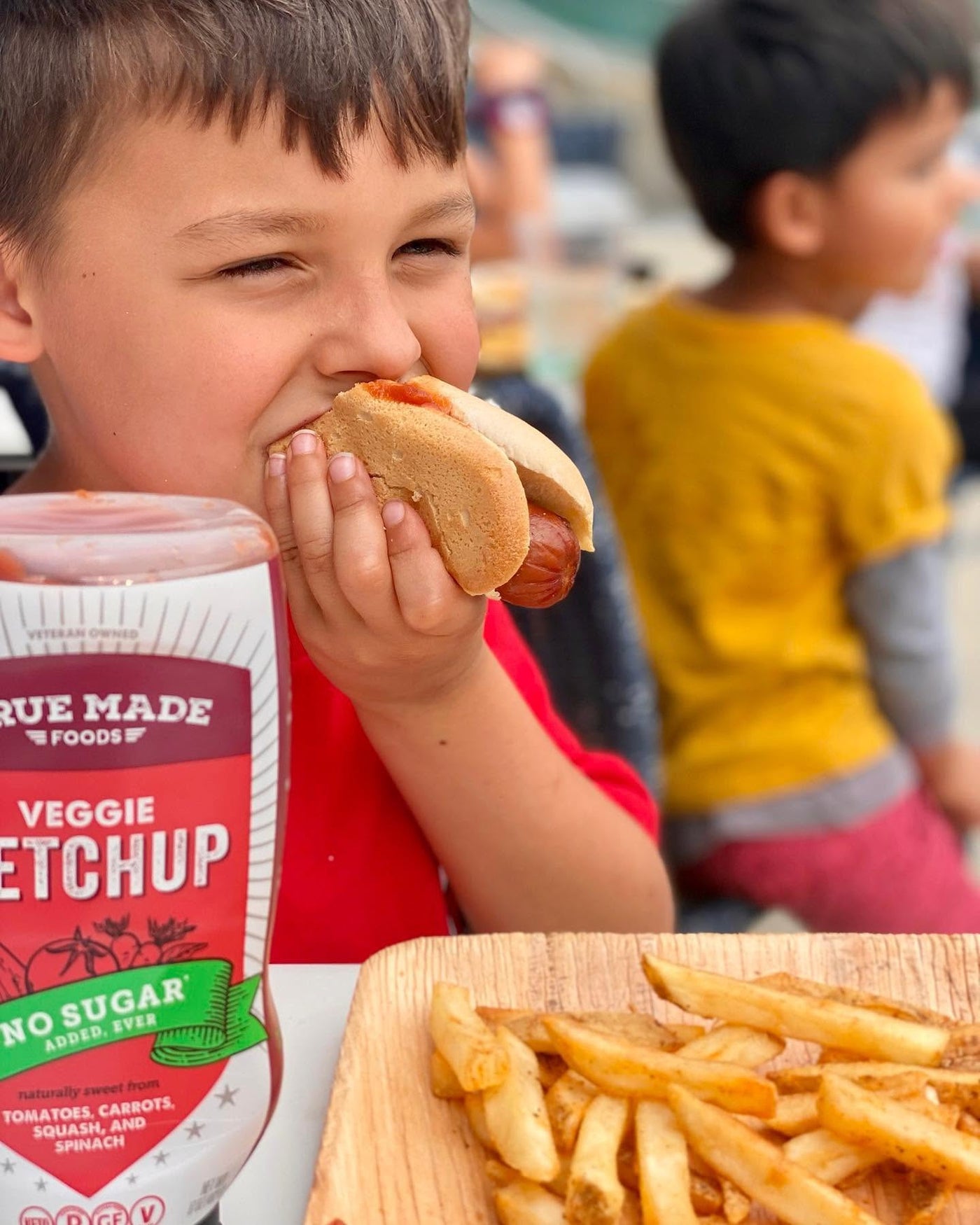 A young boy eating a hot dog with True Made Foods sugar-free veggie ketchup and a side of fries.
